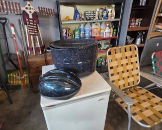 Compact white upright freezer, a large black speckled graniteware canning pot with lid, a blue bicycle helmet, and a woven-webbed aluminum folding lawn chair, with shelves of lawn chemicals, automotive fluids, and cleaning supplies plus a folk art wooden windmill in the background.
