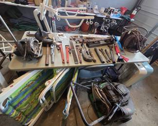 Folding table of plumbing and mechanic tools including Ridgid-style pipe wrenches in several sizes, a pipe cutter, pipe threader dies, large open-end wrenches, files, a Black and Decker circular saw, and a vintage air compressor unit; folding aluminum lawn chairs and a gas push mower below.