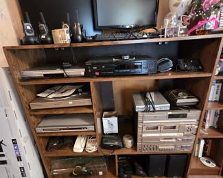 Shelf unit of home electronics featuring a Sony compact disc player, a Panasonic DVD player, a Marantz component stereo stack with CD player, dual cassette decks, and equalizer, KLH bookshelf speakers, Motorola Talkabout two-way radios, a flat-panel computer monitor, and a milk glass table lamp.