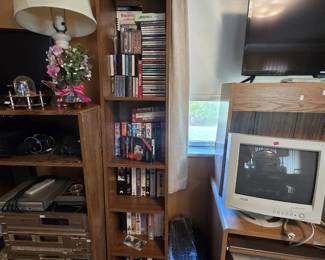 Tall media tower stocked with cassette tapes, CDs, and VHS movies including classic titles, a cassette demagnetizer, and a video head cleaning kit; nearby are a KDS CRT computer monitor, a vintage beige desktop PC tower, a flat-screen monitor, and a FAMILY wall plaque.