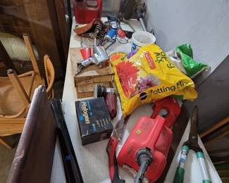 Work table with a Chicago Pneumatic air impact wrench, vintage spring-suspension bicycle saddle, red plastic gas cans, Craftsman string trimmer, Miracle-Gro potting mix, Sylvania H4656 halogen headlight in box, pruning saw, hedge shears, and a large kitchen knife, with bentwood chairs and a wood drop-leaf table nearby.