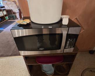 Oster countertop microwave with stainless trim and black cabinet, topped by a large vintage white enamelware stock pot with black rim and side handles. A two-shelf wood stand below holds serving trays and an aqua enamel pan.