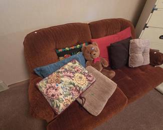 Rust orange chenille loveseat with attached pillow-back cushions, shown with an assortment of throw pillows including a floral tapestry pillow with cord trim, a needlepoint fruit pillow, and solid accent pillows, plus a large plush teddy bear with red bow tie.