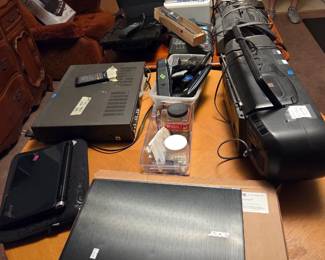 Table with electronics including an Acer laptop, a DVD or VHS player, and assorted small devices.