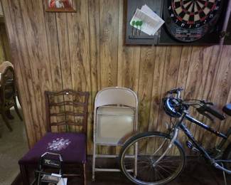 Wall grouping with an electronic dartboard in a black wall-mount cabinet with darts, a framed print of Pope John Paul II in an oak frame, a mahogany ladder-back side chair with purple needlepoint floral seat, a metal folding chair, a Travel Caddy folding luggage cart, and a mens multi-speed bicycle.
