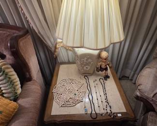 French Provincial end table with inset stone top holding a ceramic table lamp with raised floral branch relief and scalloped pleated shade, a vintage kewpie-style boxing doll, a shell trivet, and several strands of vintage crystal and jet-black beaded necklaces.