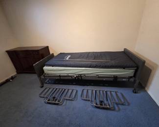 Drive Medical electric hospital bed with adjustable frame, remote control, innerspring mattress, and Drive pressure-relief mattress topper, complete with removable side rails. A mahogany server cabinet with carved doors stands alongside.