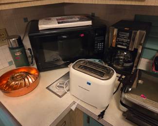 Small kitchen appliances including a microwave, coffee maker, and toaster, with a copper mold and a green thermos nearby.