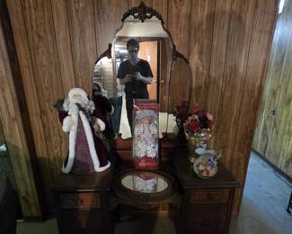 Antique 1930s vanity with carved shield-shaped mirror, two banks of drawers, and turned legs on casters, displayed with a Century Collection porcelain doll in original box, a Santa figure, a gold-toned vase with roses, and a hand-painted fruit-motif pitcher.