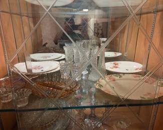 Middle shelf of the same cabinet with floral-pattern china plates, glass stemware, and amber depression glass.