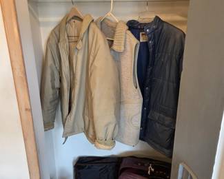 Closet of men's outerwear including a tan hooded work jacket, knit sweater vest, and navy quilted snap-front coat, with straw sun hats and a red Club Filarets trucker cap on the shelf above. Burgundy soft-side luggage, a garment bag, work boots, and shoes fill the floor below.