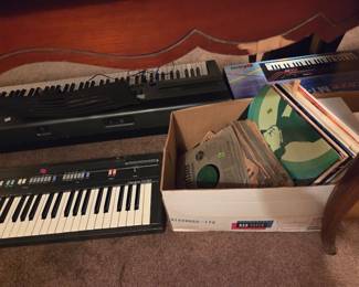 Additional electronic keyboards stacked together, next to a box of vinyl records.