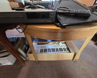 Vintage Casio Casiotone CT-102 keyboard in its original box, shown alongside a Casiotone MT-240 box, a digital piano, and a collection of vinyl records beneath a wood demilune console table. The tabletop holds a Sharp VC-A206 VHS player, a compact DVD player, a flat screen TV, and assorted remotes.