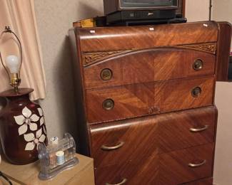 Art deco style wood highboy dresser with chevron-patterned drawers, topped with a small television and shown beside a floral hurricane lamp.