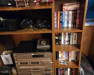 Lower section of the same shelf with a stereo cassette component, small speakers, and more VHS movies.