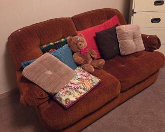 Rust-colored plush loveseat with assorted throw pillows and a teddy bear, next to a set of white stacking storage drawers.