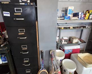 Five-drawer black metal vertical file cabinet with a shelf stereo system on top, vintage Spalding wooden tennis racquet, wooden badminton racquets, a Rubbermaid 48-quart cooler, coolers, and household supplies on garage shelving.