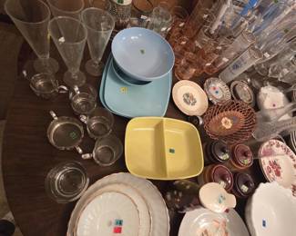 Table of vintage glassware and dishware including tall pilsner glasses, pink-tinted tumblers, mid-century melamine dinnerware in blue, turquoise, and chartreuse, a divided serving dish, milk glass with gold trim, stoneware condiment jars, a copper wire basket, and glass cups in pierced metal holders.
