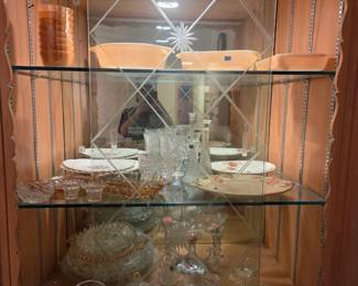 Lighted wood curio cabinet with glass shelves, displaying pink glassware, china plates, and cut glass pieces.