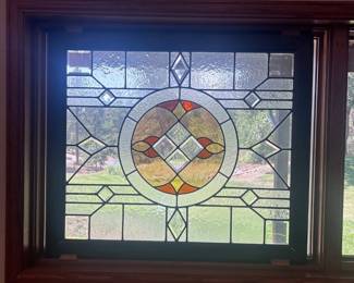 af- Large decorative stained and leaded glass window panel featuring a symmetrical geometric design with a central circular motif. Constructed with a variety of clear textured glass, amber, red and yellow-green glass accents, along with multiple beveled glass pieces that catch and reflect the light. The panel is set within a dark wood frame. Measures approximately 41.5” wide x 36” tall x 1” deep overall. There is a small crack in one glass panel that has been sealed/repaired, as shown in the photos. Otherwise, the piece presents nicely with attractive variation in the textured and beveled glass. Please review photos for condition and details.