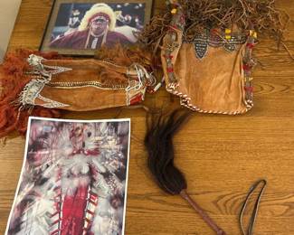 kp- This unique American Indian decor collection includes a variety of handcrafted items such as an intricately designed leather pouch adorned with detailed beadwork and fringe, a traditional feathered ceremonial accessory, and a striking printed artwork depicting a Native American figure in vibrant red and multicolored traditional attire. The set showcases rich earth tones in the leather pieces combined with natural feathers and colorful geometric patterns, highlighting the cultural craftsmanship and heritage. The largest artwork measures approximately 24 inches in height, enhancing any space with its vivid and dynamic portrayal of Native American culture.