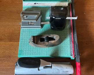 lq- This office set features a variety of essential desk tools including a sturdy paper cutter with a turquoise base and red safety handle, a black and gray dual hole puncher, a large gray hole puncher with a black rubber grip, and a unique wooden styled tape dispenser. Each item is crafted from durable materials combining metal and plastic showcasing a mix of classic design and functional utility. The set is compact, perfect for adding a retro touch to any workspace while providing practical office solutions.