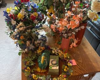 gc- A collection of beautiful floral arrangements featuring a variety of colorful artificial flowers meticulously arranged in different decorative containers. Highlights include vibrant lilies, daisies, roses, and other blooms in shades of orange, white, blue, red, and yellow. The arrangements come in diverse vases and containers including tall coral-colored ceramic vases, a golden metallic pot, and a rustic woven basket. Each piece adds a charming touch of natural elegance to any room with its detailed craftsmanship and vibrant colors.