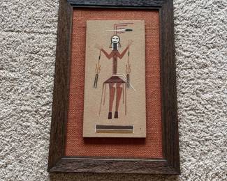 dp- Vintage framed sand art depicting a stylized ceremonial figure in traditional Southwestern-inspired colors and geometric forms. The image is created on a textured sand-like surface in shades of rust, brown, black and white and is mounted over a woven rust-colored fabric backing in a rustic wood frame. A handwritten note retained on the reverse identifies the subject as “Holy Heal People” and references the figures being used in connection with singing and healing. The exact cultural origin and artist are not identified on the piece, so no specific tribal attribution is being made. The reverse also retains a Kemper Galleries, Colorado Springs, Colorado label. The gallery label advertises original oil paintings and custom framing An attractive vintage Southwestern decorative piece with interesting handwritten provenance retained on the back.