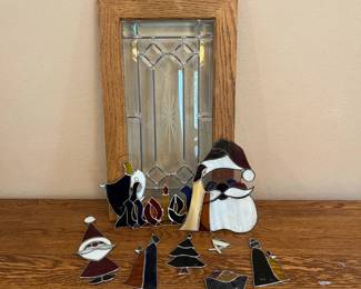 mq- This set features a collection of stained glass Christmas decorations depicting a nativity scene, Santa figures, a Christmas tree, a sleigh, and the word 'Noel'. Each piece is crafted from colorful stained glass with black soldered outlines, offering a festive and artistic appeal. The set also includes a beautiful oak framed leaded glass panel with clear textured glass and a starburst design at its center, showcasing exquisite craftsmanship. Highlighting rich colors such as deep reds, whites, blacks, yellows, and blues, along with natural oak wood tones, this collection is perfect for seasonal décor or collectors of vintage holiday items.