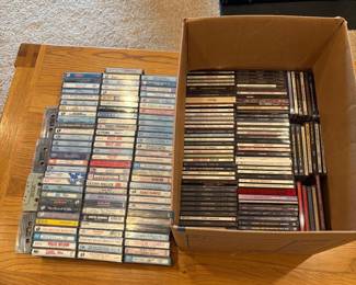 ig- This collection features a diverse assortment of compact discs and cassette tapes, neatly organized and stored in a cardboard box. The assortment includes various artists and genres, with cassettes showcasing vintage designs and labels. Highlighted aspects include the classic TDK cassette cases, a mix of color schemes on the CD spines, and a substantial size indicating a large variety of music selections perfect for collectors or enthusiasts of physical music media.