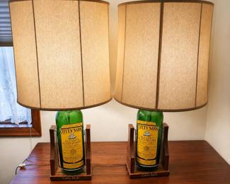 ai- This unique set features two vintage Cutty Sark Blended Scots Whisky bottles repurposed as stylish table lamps. Each lamp includes a 17-inch tall bottle with authentic yellow and green Cutty Sark labels, mounted on a custom wooden base engraved with 'Cutty Sark.' The lamps stand 34 inches tall in total, with a large 17.5-inch wide fabric shade providing a warm and inviting glow. These eye-catching lamps combine nostalgic whisky branding with functional lighting, perfect for collectors or enthusiasts seeking distinctive home décor.