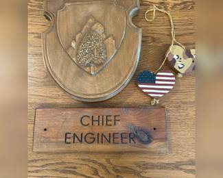 fv- This unique wooden wall decor set includes a finely carved wooden plaque with an intricately detailed tree emblem and a separate wooden sign engraved with 'CHIEF ENGINEER.' The decor pieces showcase a rich, natural wood finish highlighting the beautiful grain and knots, adding a rustic and authentic touch. Additionally, the set is complemented by two charming heart-shaped wooden ornaments, one painted with the American flag and the other in a camouflage pattern, each hung with rustic twine. Perfect for adding character and a personalized feel to any room or office space.