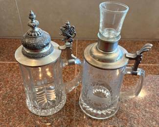 lja- This German style beer stein set features two unique glass steins adorned with intricately detailed pewter lids and lion-themed thumb lifts. The set includes a 9-inch tall stein with an ornate lion thumb lift, a 7.5-inch stein without a shot glass, and a 9.25-inch stein with an integrated shot glass on top. Both steins showcase clear glass bodies with decorative patterns and finely crafted pewter accents, and one marked as made in Germany. This elegant set is perfect for collectors or enthusiasts of traditional German beerware, highlighting fine metalwork, distinctive lion motifs, and varying sizes to suit different serving preferences.