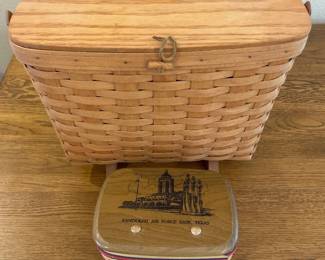 fs- This set features two unique Longaberger baskets known for their handcrafted quality and classic woven wood design. The larger basket showcases a natural wood finish with a sturdy latch and is elevated on small wooden feet. Accompanying it is a smaller, patriotic-themed basket adorned with red, white, and blue bands and topped with a Randall Air Force Base, Texas commemorative tin lid, highlighting a historical military base illustration. Both pieces emphasize traditional craftsmanship, durable wood material, and distinctive designs suitable for collectors and basket enthusiasts.