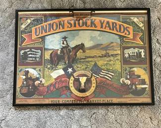 ec- Framed commemorative poster celebrating 100 years of the Union Stock Yards in San Antonio, Texas, 1889–1989. The colorful Western-themed artwork features a mounted cowboy, Texas and American flags, longhorn imagery, a steam locomotive, semi truck, and historic buildings, along with the wording “Serving the Southwest,” “100th Anniversary,” and “Your Competitive Market Place.” The bottom border lists several livestock commission firms and organizations associated with the stockyards, including J.W. Kothmann & Sons Live Stock Commission Agents, Alamo Livestock Commission Company, Producers Live Stock Commission Agents, Texas Livestock Marketing Association, and Union Live Stock Commission Company. This appears to be the poster designed by Thom Ricks commemorating the San Antonio Union Stock Yards centennial.