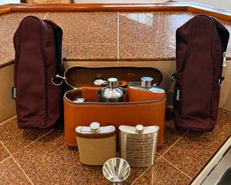 ld- This vintage travel bar set features a stylish brown leather case containing multiple stainless steel flasks and a portable wine carrier. Included are two flasks, one with a fabric-covered exterior and another engraved with detailed text, along with a small metal funnel for easy pouring. The set also comes with two dark brown zippered wine carrying bags enhancing portability and protection. Perfect for travel or outdoor enjoyment, this set combines classic design with practical utility.