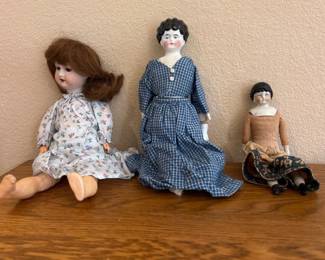 kw- This charming set includes three antique ceramic dolls, each with unique hand-painted facial features and distinct clothing styles. The dolls vary in size, with the largest measuring approximately 14 inches and dressed in a delicate, floral-patterned dress in soft pastel colors. The medium doll sports a blue checkered dress with buttons, adding to its vintage appeal. The smallest doll is adorned in a brown and black outfit, showcasing intricate craftsmanship. All dolls feature ceramic heads, arms, and legs, with cloth bodies, embodying classic antique doll-making techniques. These collectible dolls make a delightful addition to any vintage toy collection or decorative display.