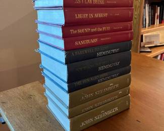 lz- This collection features eleven hardcover books showcasing works by William Faulkner, Ernest Hemingway, and John Steinbeck. The set is distinguished by its tri-color scheme: deep red for Faulkner's titles, navy blue for Hemingway's classics, and olive green for Steinbeck's novels. Each book has gold embossed titles on the spine and cover, adding a vintage elegance. The books are of uniform size with durable cloth-bound covers, each reflecting slight wear consistent with age and careful use, emphasizing their authenticity and collectibility. Ideal for literary enthusiasts and collectors, this set offers a tangible connection to some of the most influential American authors of the 20th century.