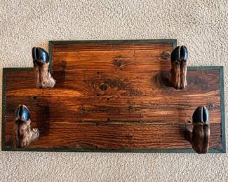 bf- Unique rustic wall-mounted rack featuring four preserved deer hooves/feet mounted to a substantial wood backing with a stepped top profile and dark green trim. The hooves project outward to function as hooks, making the piece suitable for displaying hats, coats, or other lightweight items. Photos show military hats displayed on the upper hooks for scale and use; hats are not included unless otherwise specified. The wood backing has an aged, heavily grained appearance with knots, cracks, scratches, and other character consistent with use and rustic construction. Taxidermy elements show natural variations and age-related wear. An unusual piece well suited for a hunting lodge, cabin, western décor collection, man cave, or sporting display. Approximate Dimensions: ● 22” tall ● 42.5” wide ● Projects approximately 12” from the wall Please review photos closely for condition and construction details.