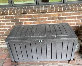 id- This outdoor storage chest is designed for convenient garden tool and supply storage. It features a durable black plastic construction with a slatted panel design and is mounted on a dolly for easy mobility. Measuring 42 inches in width and 22.5 inches in height, it offers ample space for organizing gardening essentials. The chest has a secure lid that lifts open, providing easy access to the contents inside. Its robust material ensures weather resistance, making it ideal for outdoor use on patios or porches.