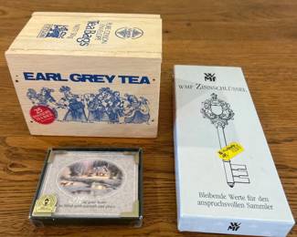 ft- This diverse collection of collector's items features a vintage wooden box of Earl Grey Tea adorned with classic blue illustrations. Included is a decorative plaque with a serene winter cabin scene and a heartfelt message, elegantly framed with gold-colored corners. Additionally, a boxed WMF pewter key is presented, showcasing intricate craftsmanship and a label highlighting its value for discerning collectors. These items vary in material, from wood and paper to pewter, and offer unique appeal to collectors and tea enthusiasts alike.
