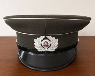 al- Original Cold War-era East German National People’s Army (NVA) visor cap, featuring a field-gray/olive body with light piping, glossy black visor and black chinstrap. The front displays the distinctive DDR national emblem with hammer and compass surrounded by a silver-tone oak-leaf wreath. Interior is clearly stamped: NVA 56 – European hat size 1856 – manufacturer code associated with VEB Perfekt Berlin, a known East German military headwear manufacturer D – production/date code The construction, markings and insignia are consistent with period East German military headwear. Manufacturer code 1856 is documented on other surviving NVA caps and has been identified with VEB Perfekt Berlin. Provenance: Acquired in Germany by the current owner, a U.S. Army battalion commander, around the time of the fall of the Berlin Wall in 1989.