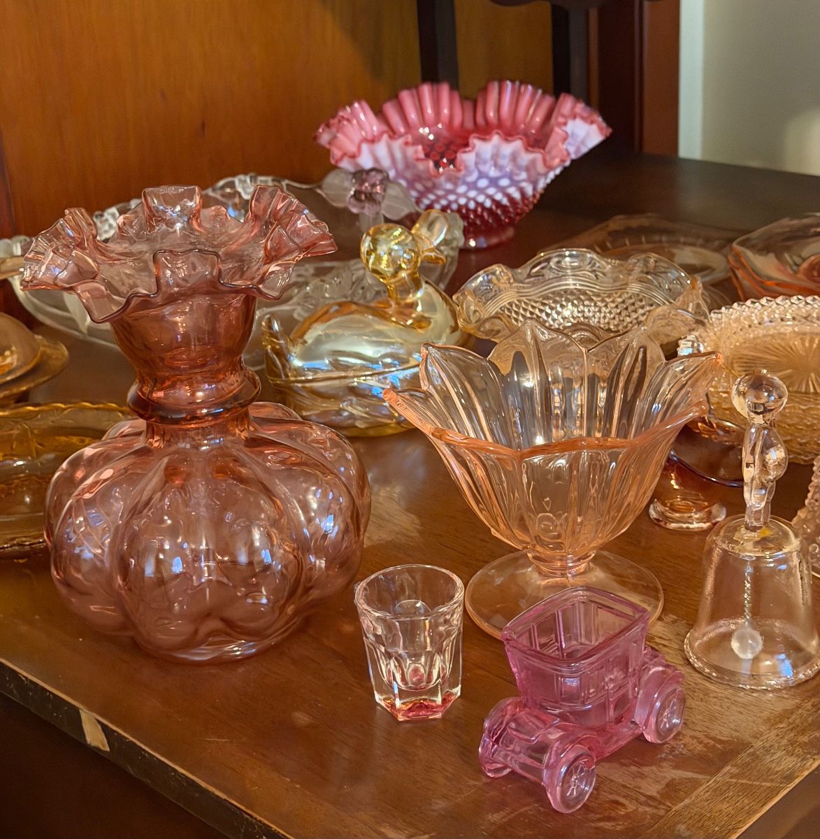 Fenton and vintage Depression glass, including a Fenton cranberry pink opalescent hobnail ruffled bowl, pink Depression glass candy compote dish, and a vintage Fenton Melon Optic cranberry vase with ruffled rim. Also includes additional pink, amber, and clear pressed-glass serving pieces, covered dishes, and decorative glassware.