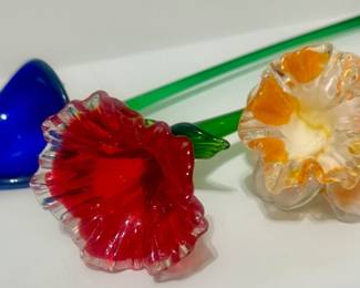 Colorful art-glass flower sculptures featuring delicate translucent green stems and vivid red, cobalt-blue, amber, and clear ruffled blooms.