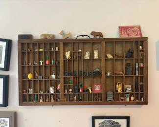 Antique wood letterpress/typesetter's type tray, wall-mounted as a curio shelf with well over 100 small compartments filled with miniature figurines and knick-knacks: carved animals, several faux-ivory/resin 'Hotei' laughing Buddha figures, miniature souvenir beer steins, cast metal and celluloid dog/animal figurines, miniature glass advertising bottles (Tabasco, Coca-Cola), a die-cast toy car, old keys, garden gnomes, small chalkware figurines, printer's letterpress type blocks spelling the family name 'ROSENBERG' in mirror-reverse (plus 'DAD' and 'MOM' blocks and fraternity/sorority letters), a small menorah, a framed vintage child's photo, miniature cast-iron stove and wringer-washer novelties, a dreidel, and more; two carved wood animal figures and a 'Southern Belle' souvenir card sit on top of the frame.