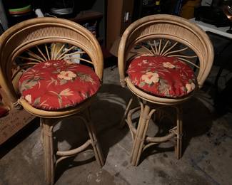 Vintage 29" swivel bar stools are constructed from natural rattan and wicker. Each stool has a curved barrel-style backrest with a decorative fan-shaped support. The circular seats are topped with removable red floral print cushions. The base is reinforced with a circular footrest and bound with rattan wraps.