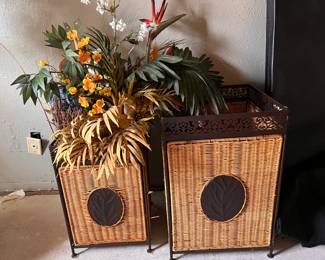 Decorative wicker and metal basket set features a woven rattan body with a dark brown metal frame. Each piece has an ornate scrollwork border at the top and a circular metal medallion with a leaf motif on the front. The baskets are supported by four small metal feet.