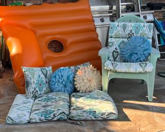 Plastic Adirondack chair in a light green finish with a matching ikat-patterned cushion set. The set includes a full chair cushion, a matching square throw pillow, and two additional seat cushions in the same teal and grey ikat print. Two decorative 3D flower pillows in blue and cream are included. (orange cheetos float not included. )