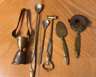 Vintage brass barware set includes a jigger, ice tongs, long stirring spoon, small spoon, bottle opener with hammer head, cheese knife, and a cocktail strainer. Each piece is made of brass with ornate decorative handles.