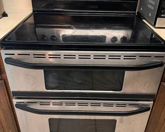 Stainless steel electric range has a double oven configuration and a black glass cooktop. This Maytag Gemini model is equipped with five heating elements including a warming center. The control panel has digital displays and tactile buttons for oven functions, while four knobs control the cooktop burners. The exterior is finished in brushed stainless steel with black handles.   Discount Note: The glass cooktop is crac